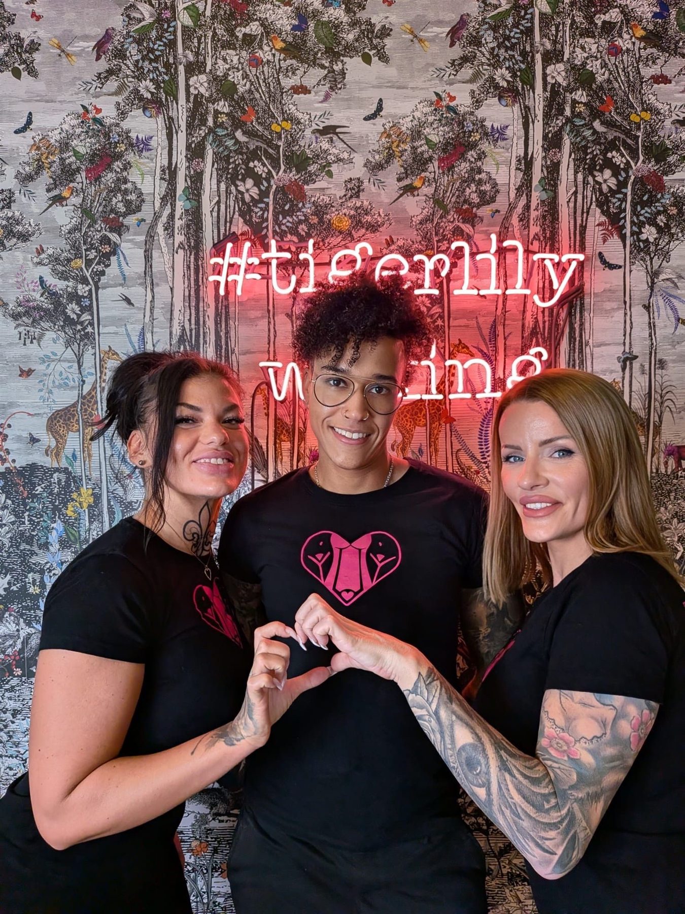 Drei Mitglieder des Tigerlily-Teams lächeln gemeinsam vor dem Neon-Schriftzug des Studios